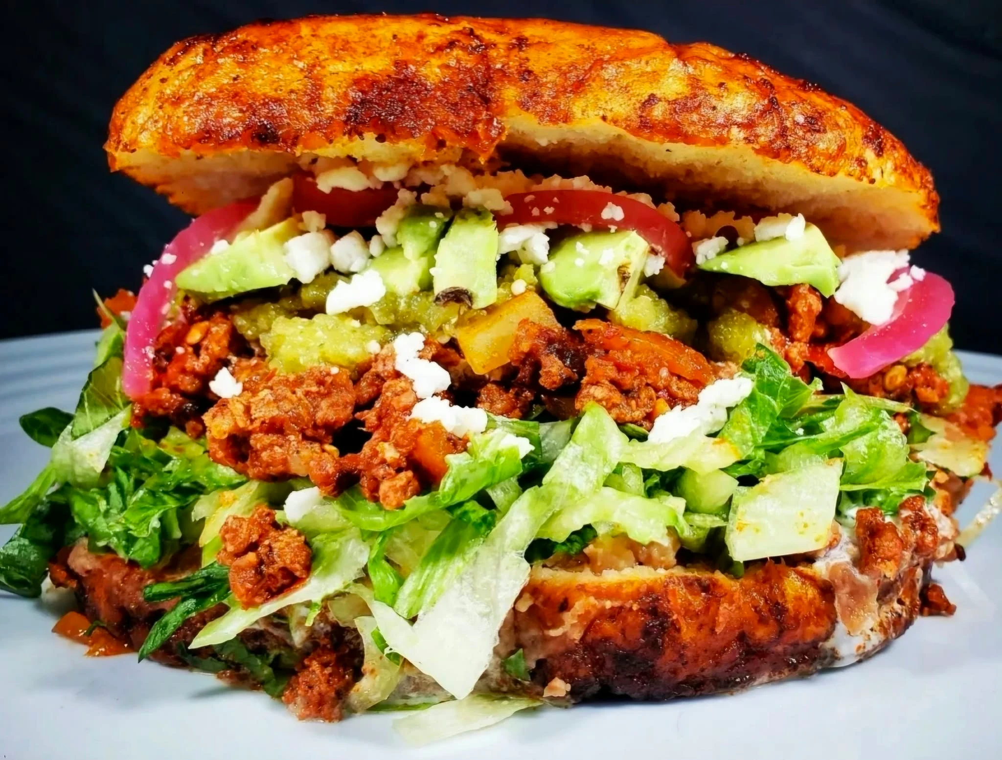 Sauce-coated pambazo sandwich filled with chorizo, potatoes, lettuce, cheese, and toppings